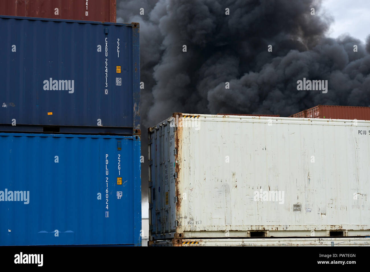 Vista astratta di contenitori di spedizione con pennacchi di fumo tossico da un incendio industriale sollevarsi verso il cielo. Foto Stock