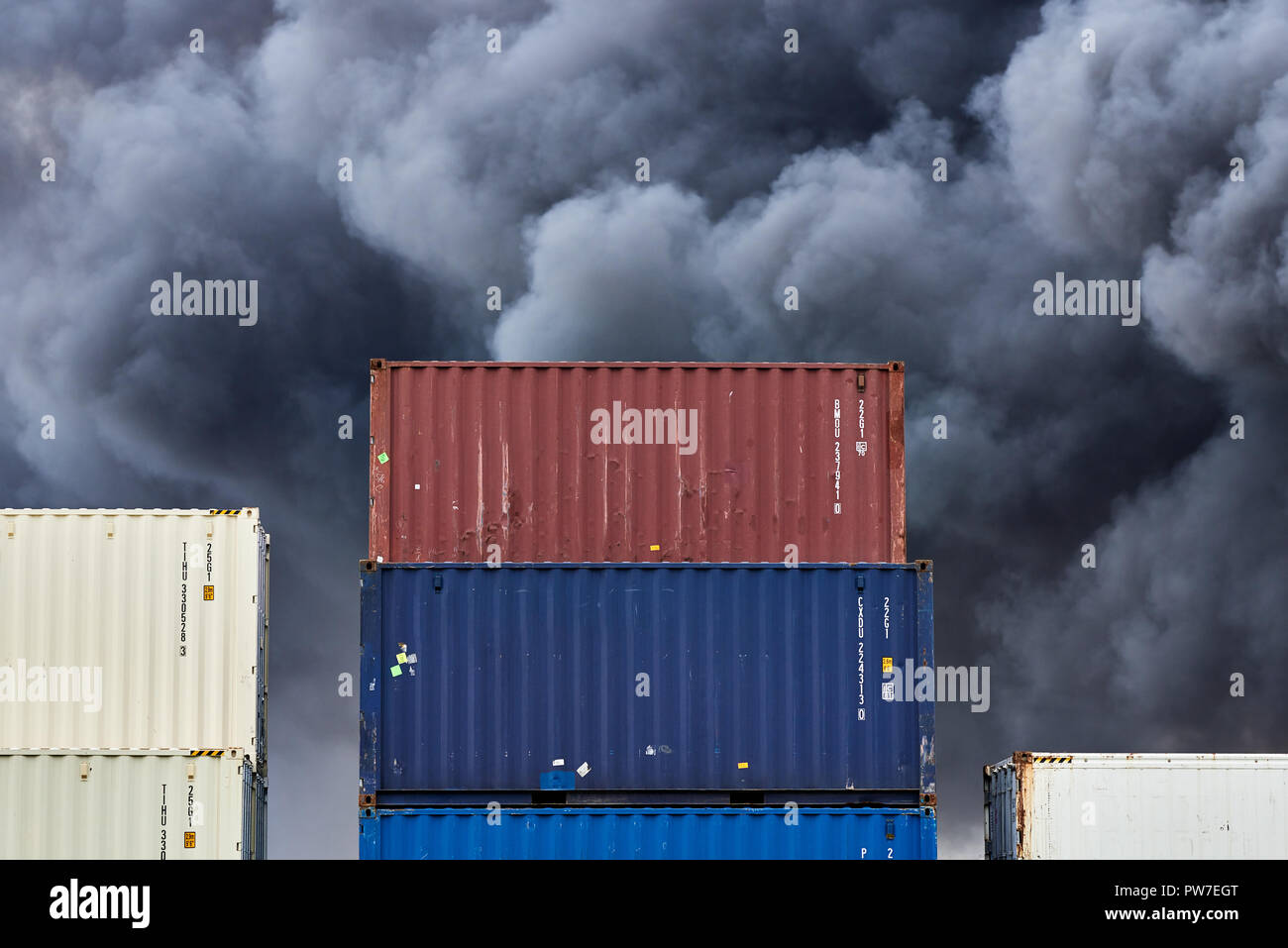 I contenitori di spedizione impilati in storage con pennacchi di nero fumo tossico da un incendio dietro. Foto Stock
