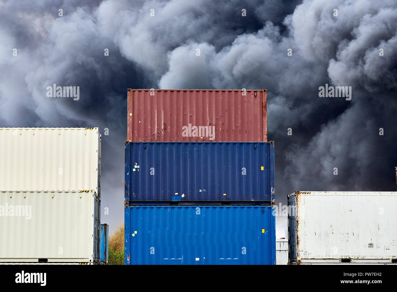 I contenitori di spedizione impilati in storage con pennacchi di nero fumo tossico da un incendio dietro. Foto Stock