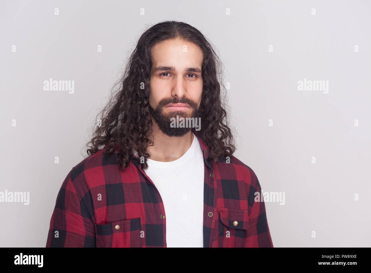 Ritratto di triste uomo bello con barba nera e lunghi capelli ricci in stile casual, a scacchi maglietta rossa in piedi e guardando la fotocamera con infelice fac Foto Stock