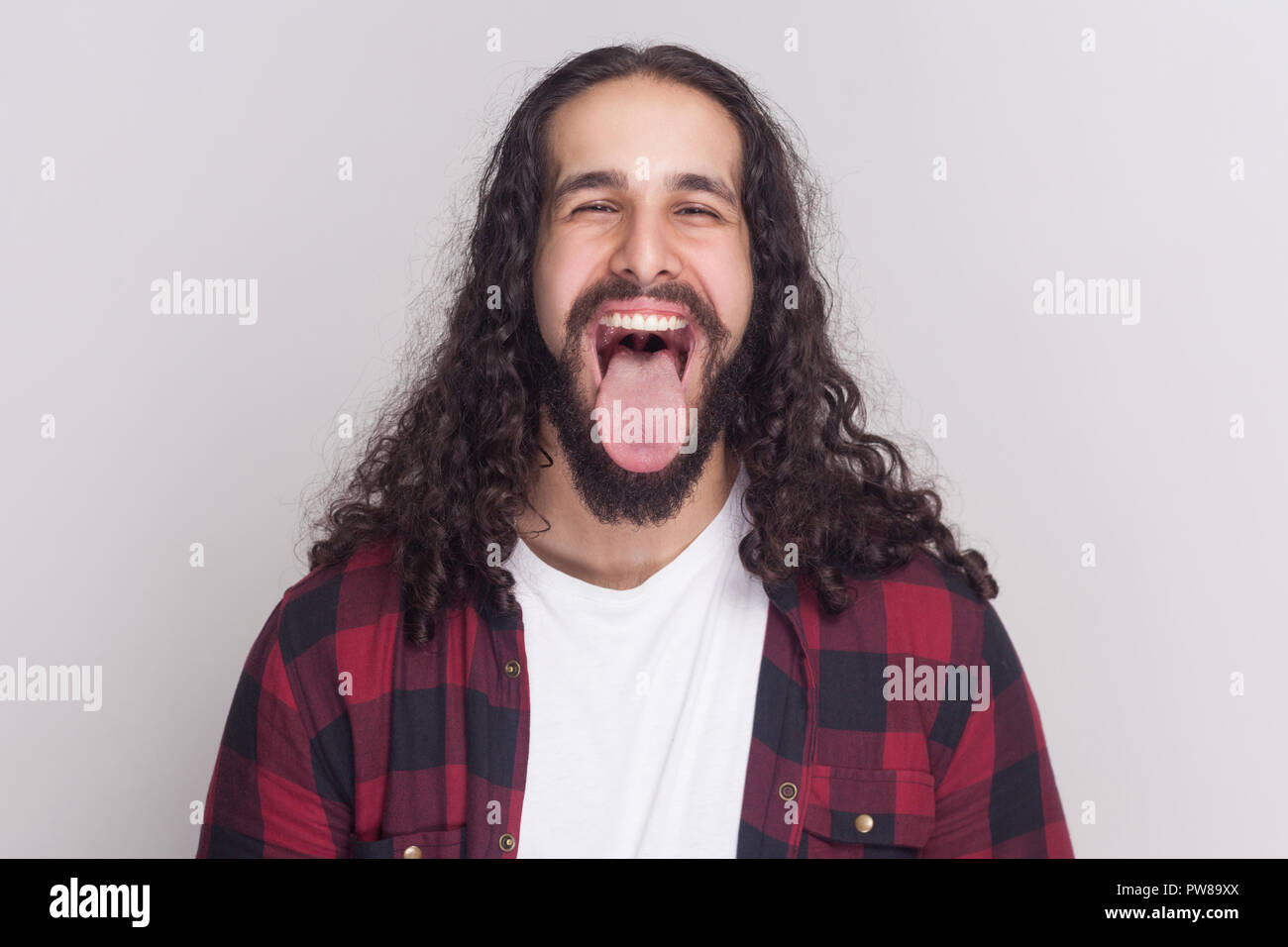 Crazy uomo bello con barba nera e lunghi capelli ricci in stile casual, a scacchi maglietta rossa in piedi, aperto bocca grande, la lingua di fuori e guardando camer Foto Stock