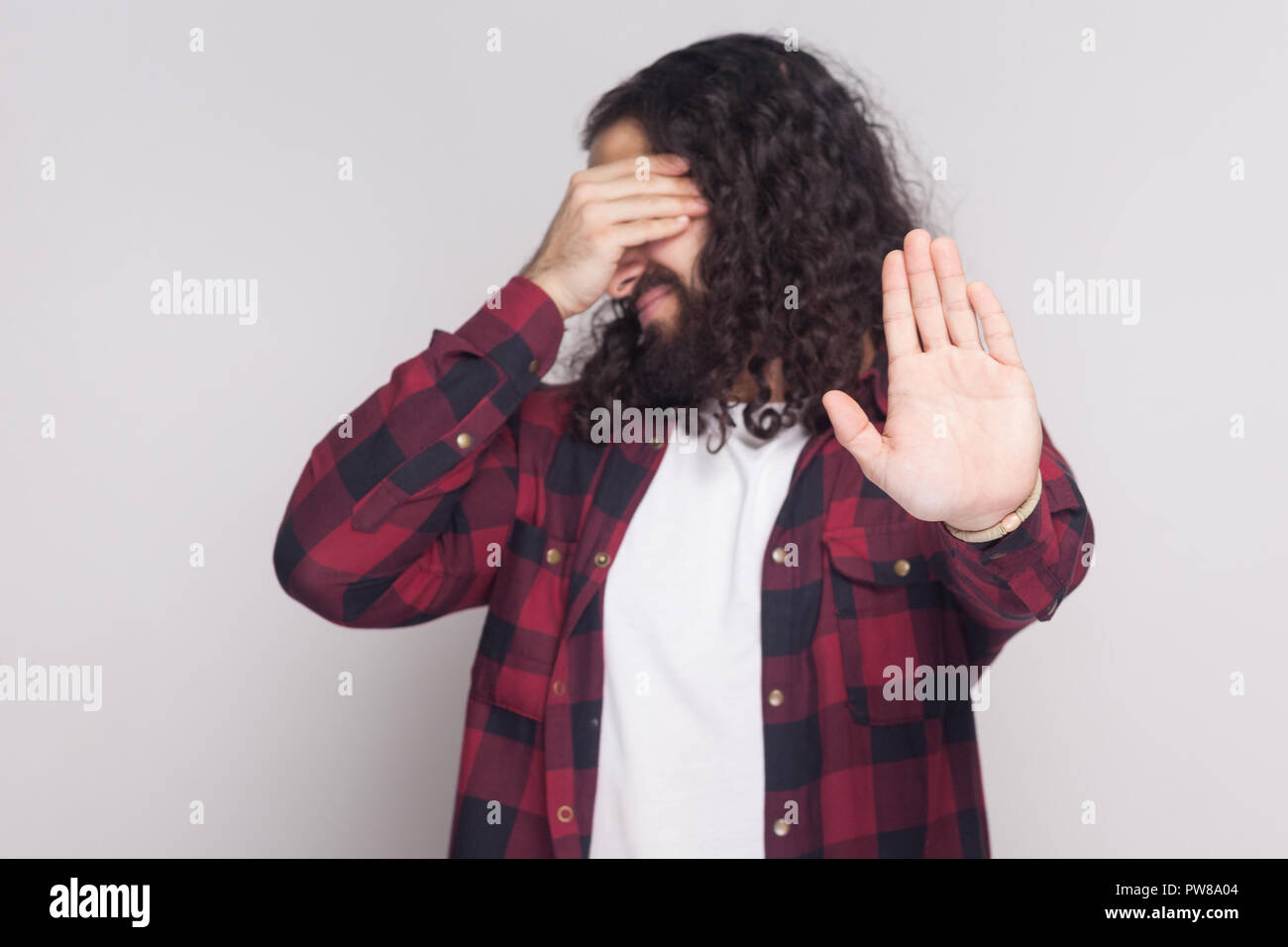 Non voglio vedere questo. Ritratto di uomo bello con barba nera e lunghi capelli ricci in rosso a scacchi camicia di copertura permanente di occhi e gesto di arresto. Foto Stock