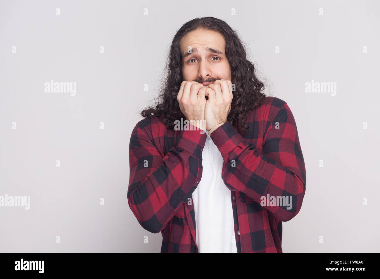 Ritratto del sistema nervoso o turbare l uomo con la barba nera e lunghi capelli ricci in stile casual, a scacchi maglietta rossa in piedi guardando la fotocamera e bitting chiodo Foto Stock