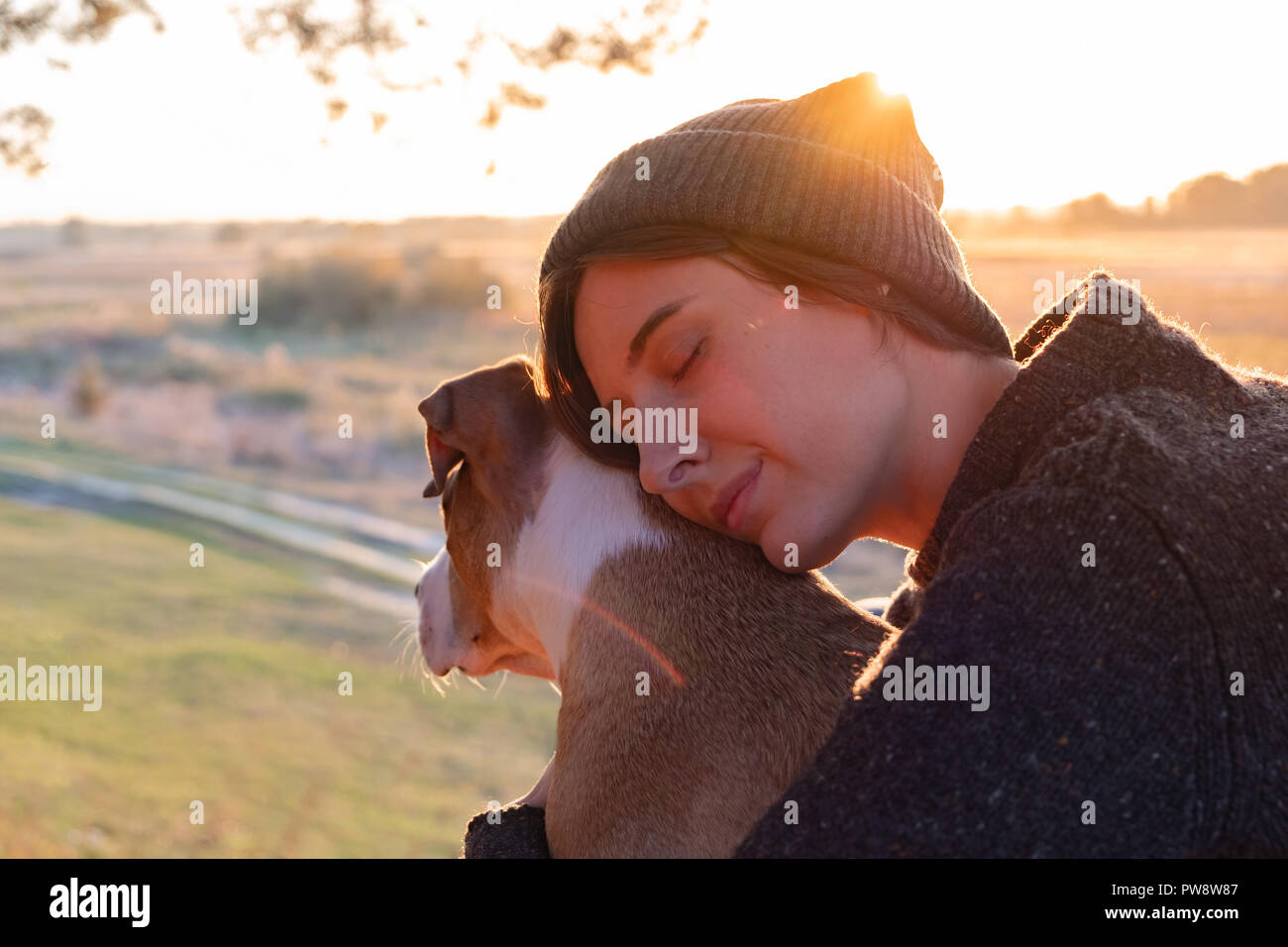Abbracciando un cane nella bellissima natura al tramonto. La donna rivolta verso il sole di sera si siede con il suo animale domestico accanto a lei Foto Stock