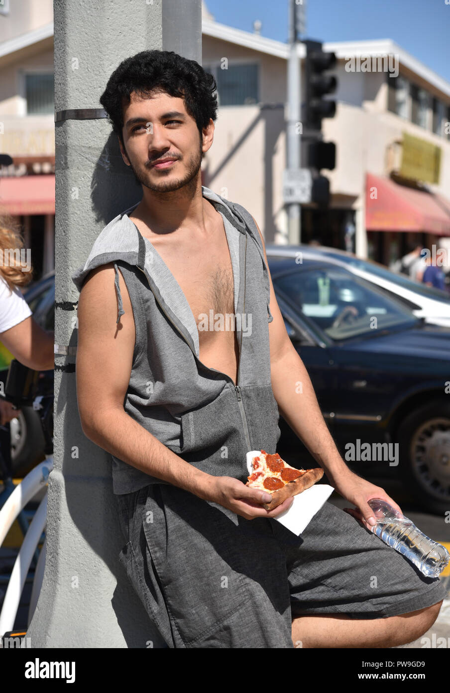 Bel giovane multi-etnico uomo appoggiato a una lampada posta in una cittadina balneare di mangiare una fetta di pizza Foto Stock