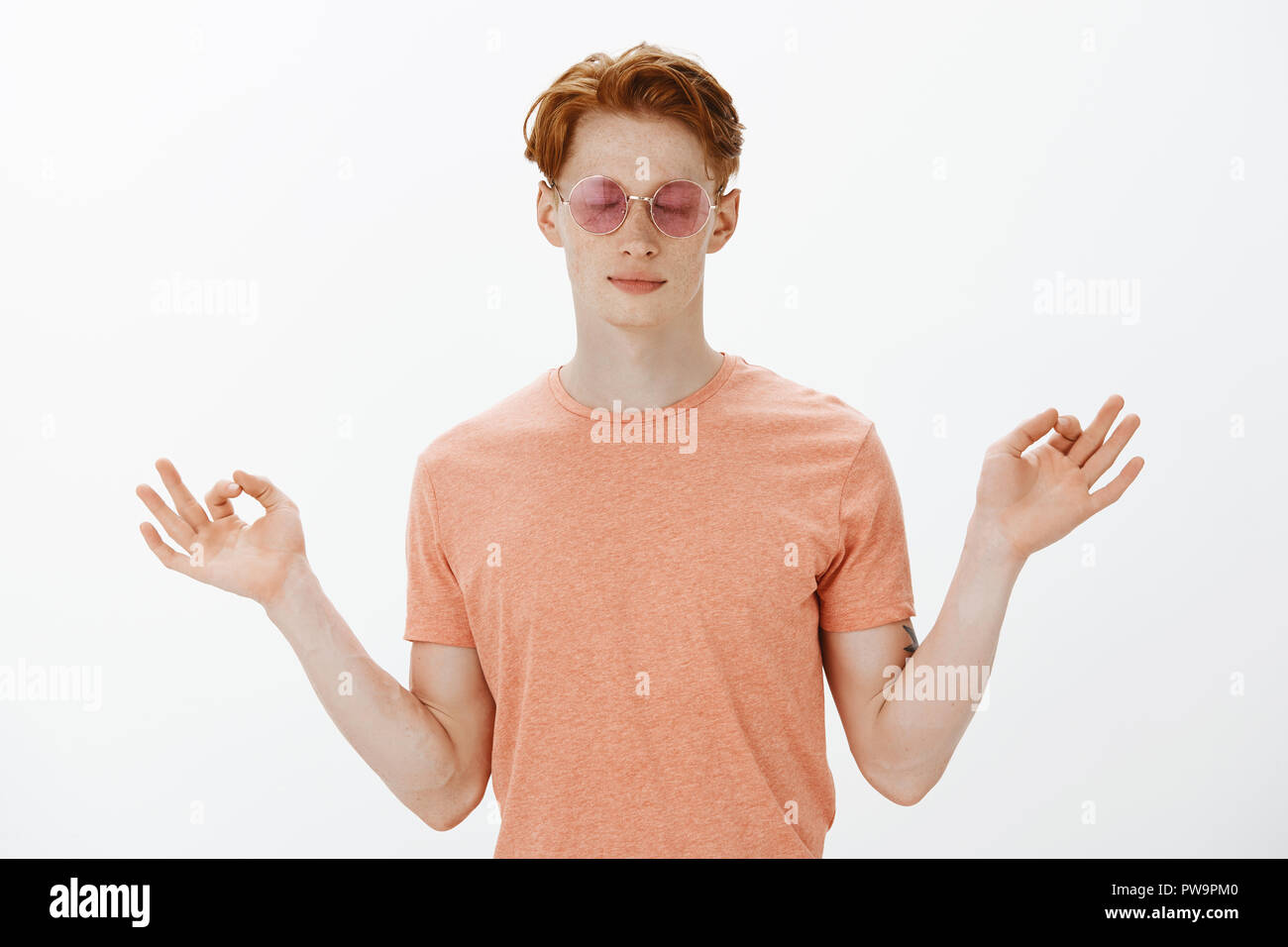 Calma e tenere il partito. Carino elegante redhead giovane uomo caucasico con lentiggini in occhiali da sole e t-shirt, chiudendo gli occhi e diffondere le mani nel gesto zen, meditando o la pratica dello yoga Foto Stock