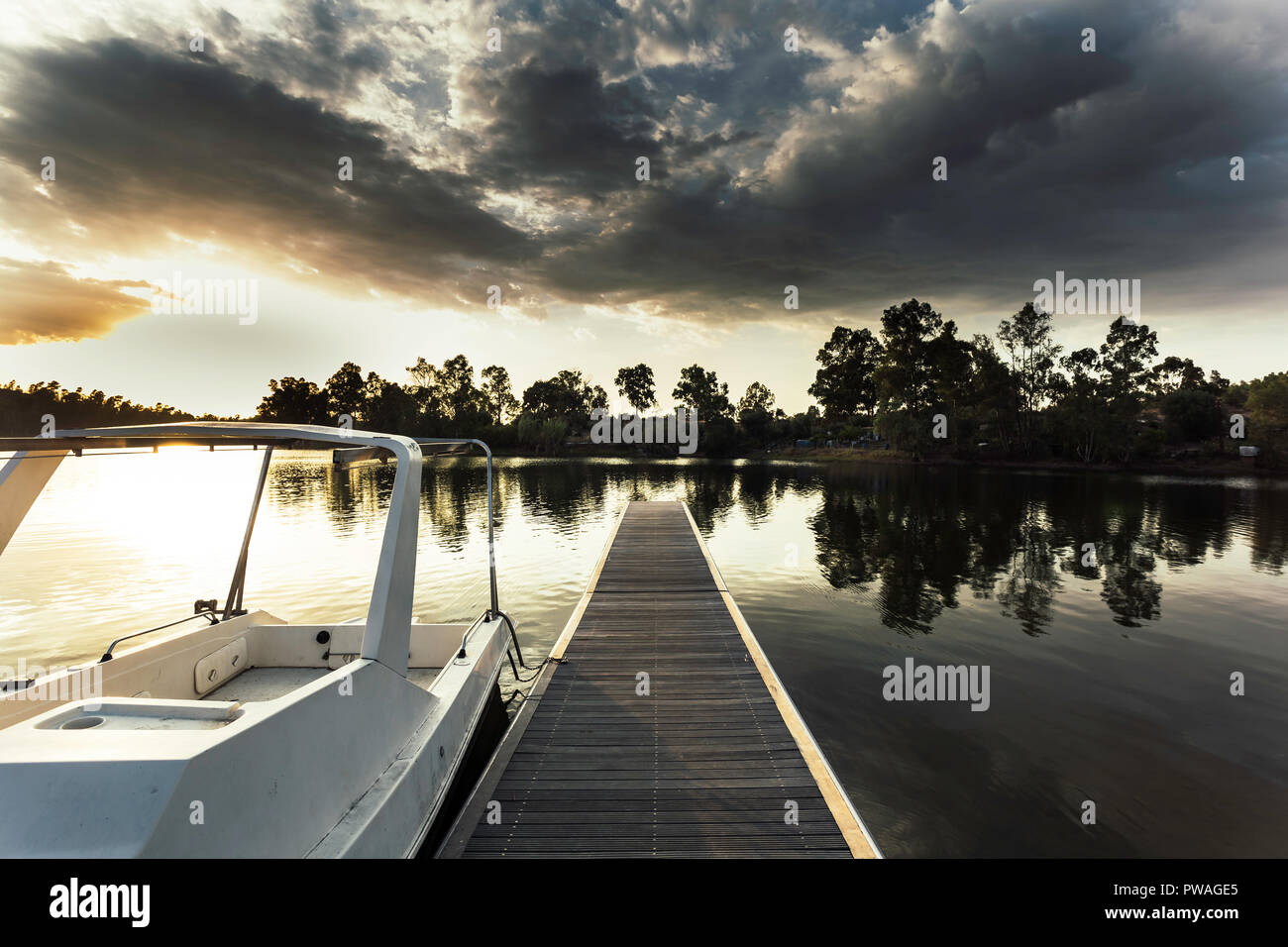 Pontile in legno con la barca ormeggiata presso la pala e riflessi dorati sulle acque del lago con un cielo coperto da nuvole in background Foto Stock