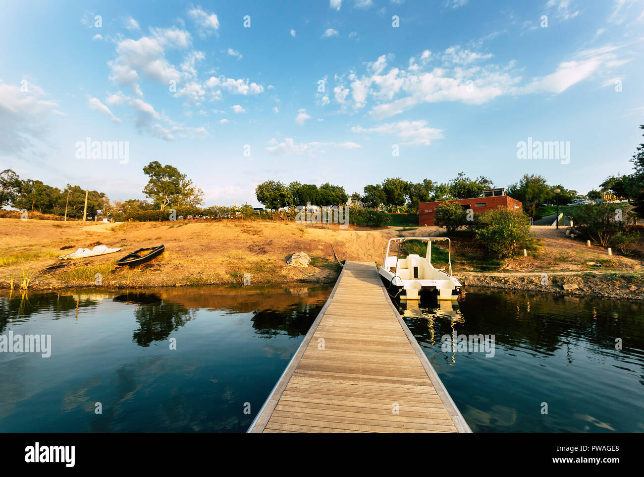 Pontile in legno con barca ormeggiata e riflessioni sul lago di acqua con un cielo blu con nuvole in background Foto Stock