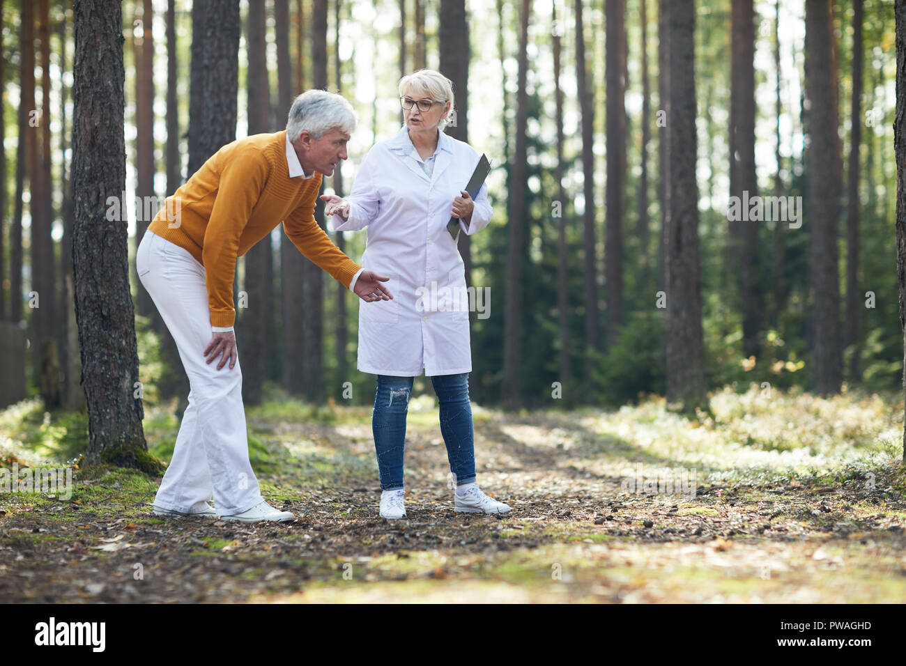 Malati senior uomo toccando il suo male la gamba mentre camminavamo lungo una strada forestale con medico aiutandolo Foto Stock