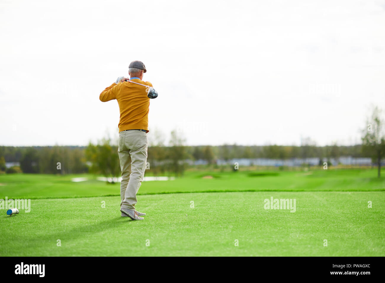 Vista posteriore del senior uomo nel cappello, maglione giallo e pantaloni beige colpire la palla con il club di golf sul campo verde Foto Stock