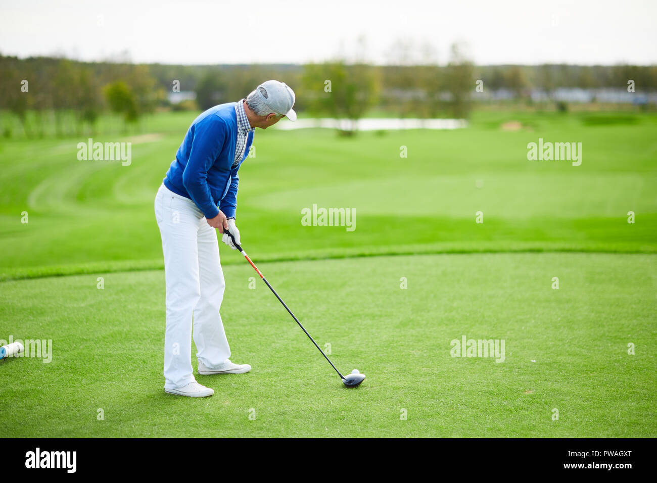 Uomo più anziano in blu cardigan, mutanda bianca e cappello da baseball preparando per colpire la pallina da golf con il club Foto Stock