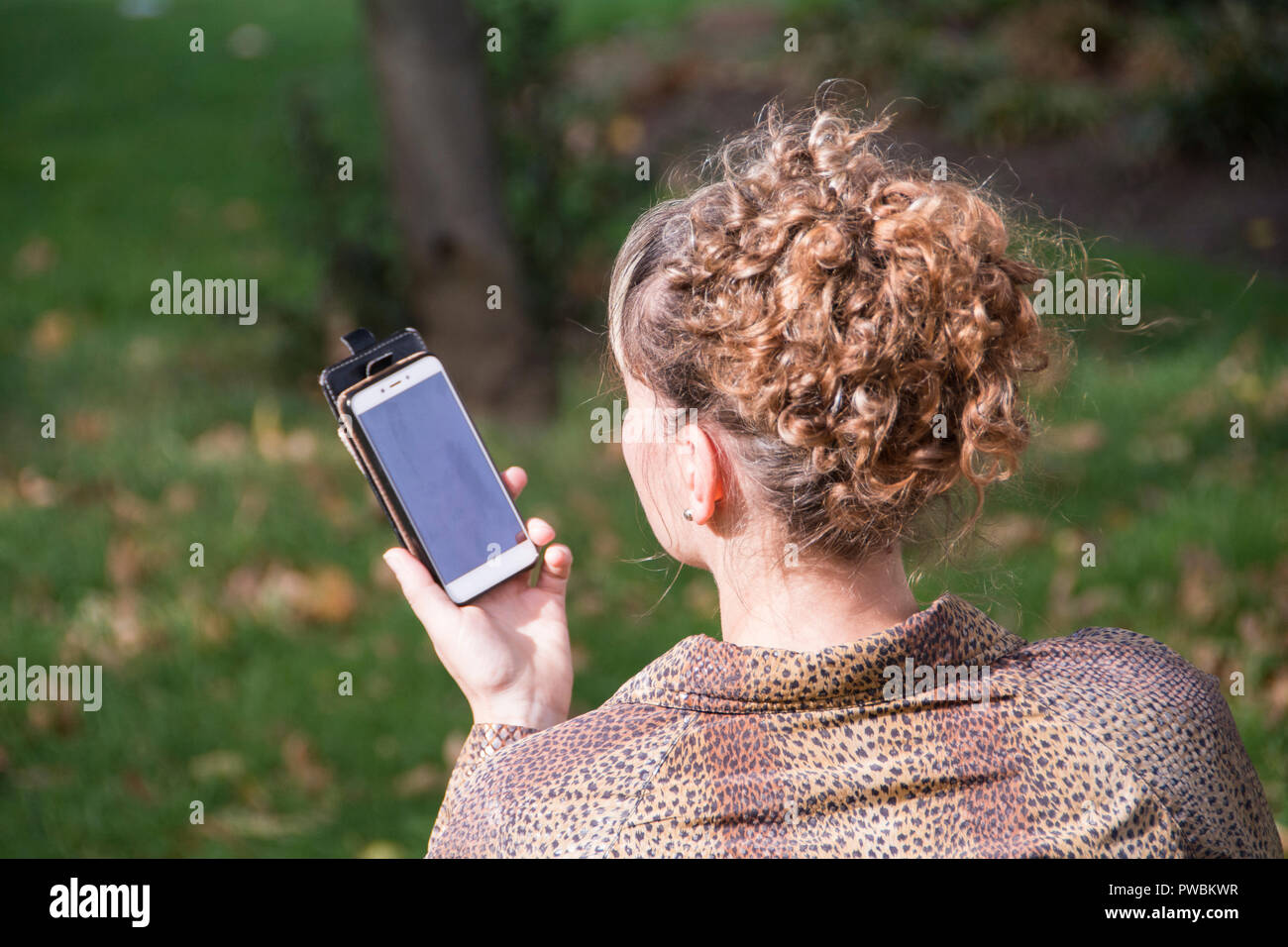 Una donna dalla testa rossa seduta da sola su una panchina del parco che effettua una telefonata sul suo cellulare a Londra, Regno Unito Foto Stock