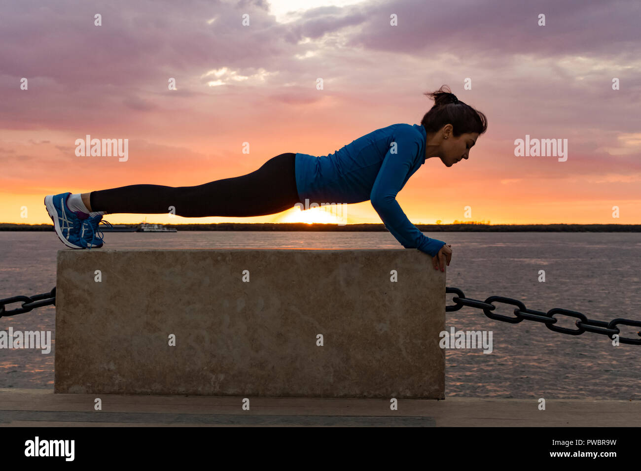 Una donna attraente in abbigliamento sportivo facendo esercizi sportivi in natura contro il tramonto e il fiume di Amur, ama la ginnastica, massaggia le gambe. Attivo giovane ragazza impegnate nello sport, conduce uno stile di vita sano. Foto Stock