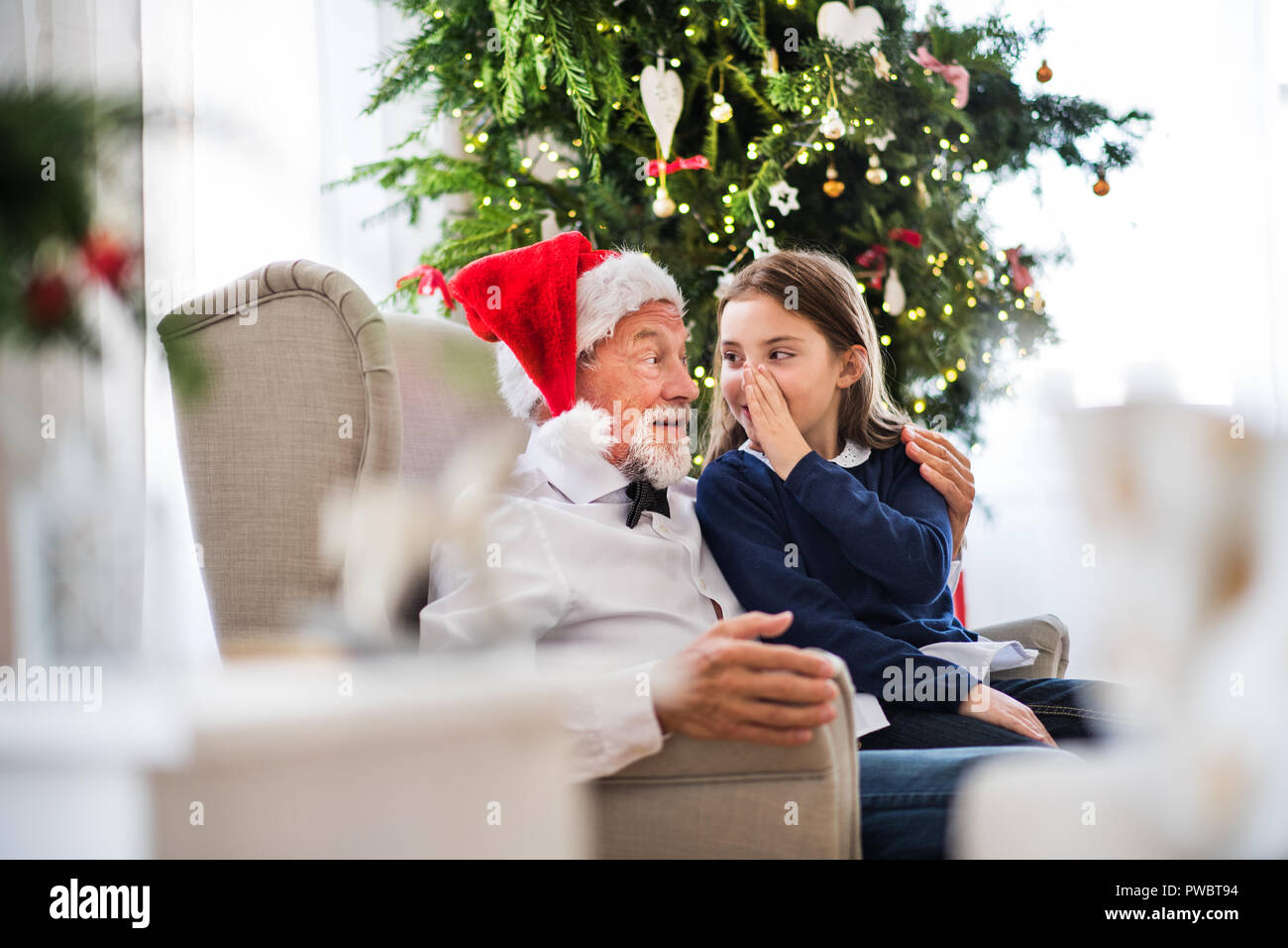 Una piccola ragazza e il nonno con Santa hat seduta sulla poltrona del tempo di Natale, raccontando un segreto. Foto Stock