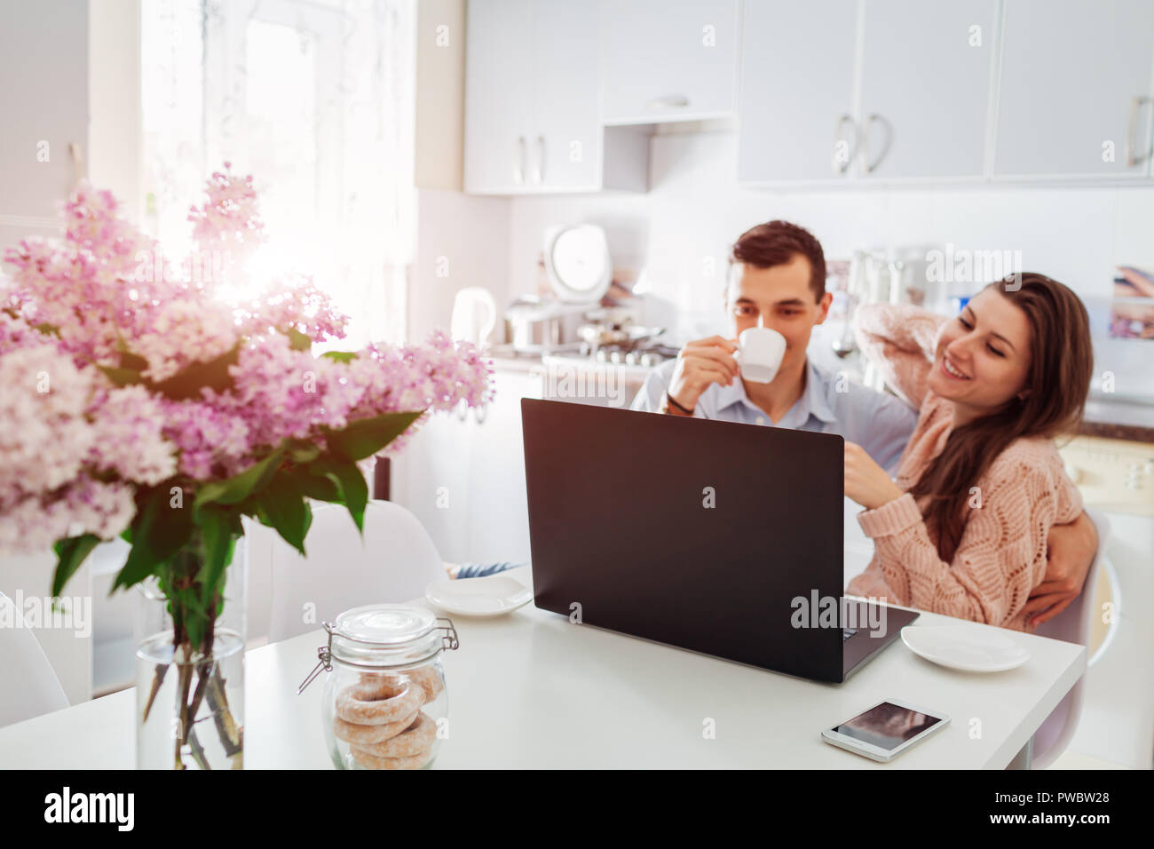 Felice coppia giovane utilizzando laptop mentre si consuma la colazione nella cucina moderna. Giovane uomo e donna bere il caffè e il sorriso. Focus su laptop Foto Stock