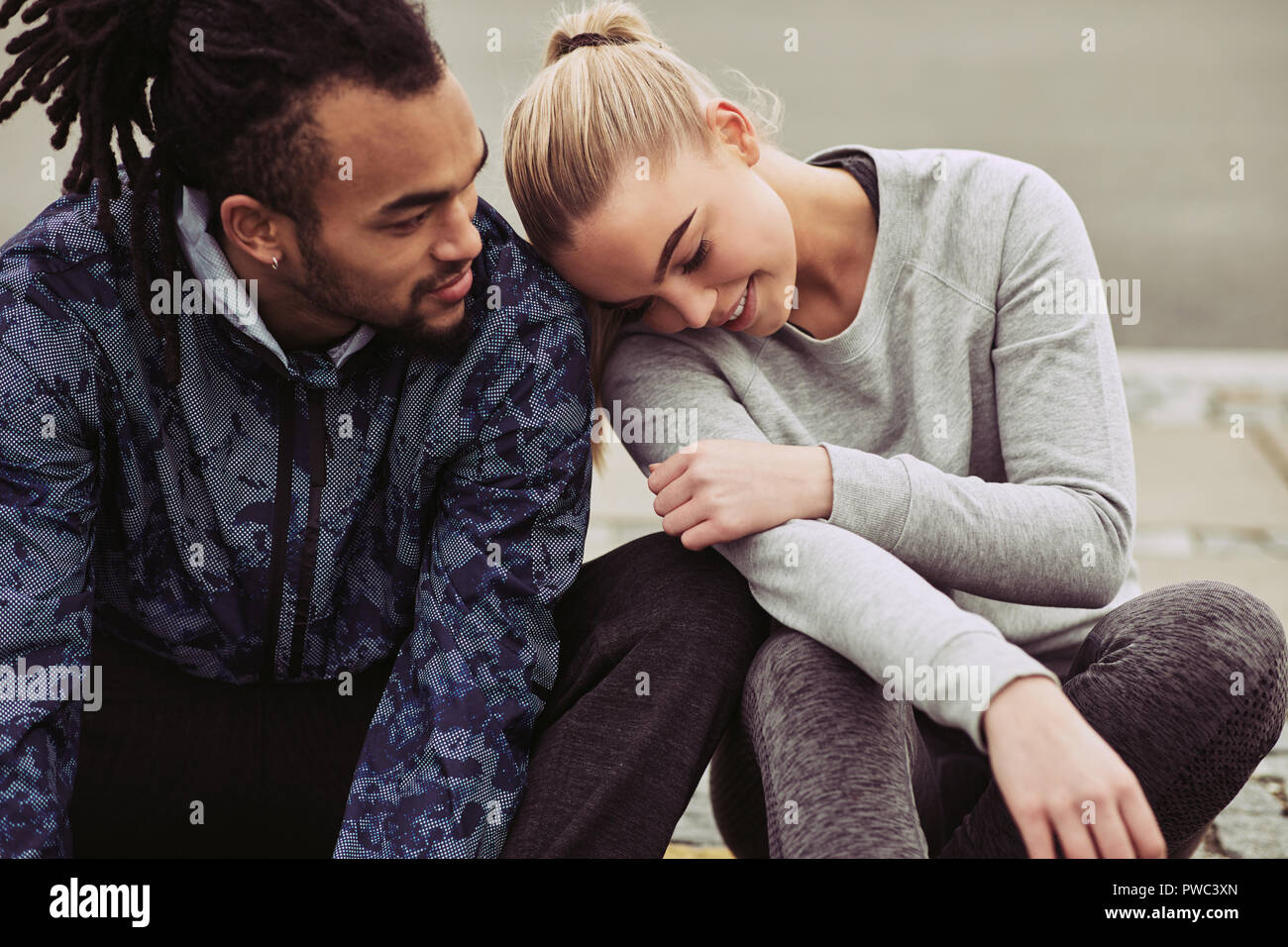 Sorridente giovane donna seduta su una strada in appoggio la sua testa sul suo fidanzato la spalla mentre prendendo una pausa insieme da un jog Foto Stock