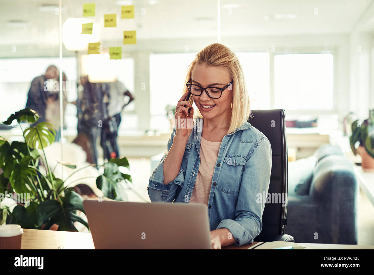 Casualmente vestito giovane imprenditrice sorridere mentre seduti ad una scrivania in un ufficio a parlare sul suo cellulare e lavorare online con un computer portatile Foto Stock