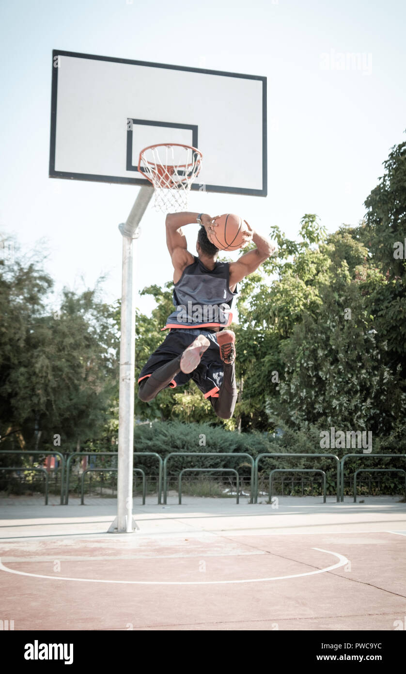 Uno afroamerican giovane uomo è giocare a basket in un parco a Madrid durante il periodo estivo a mezzogiorno. Egli sta facendo un salto impressionante per fare un dunk e mettere il b Foto Stock