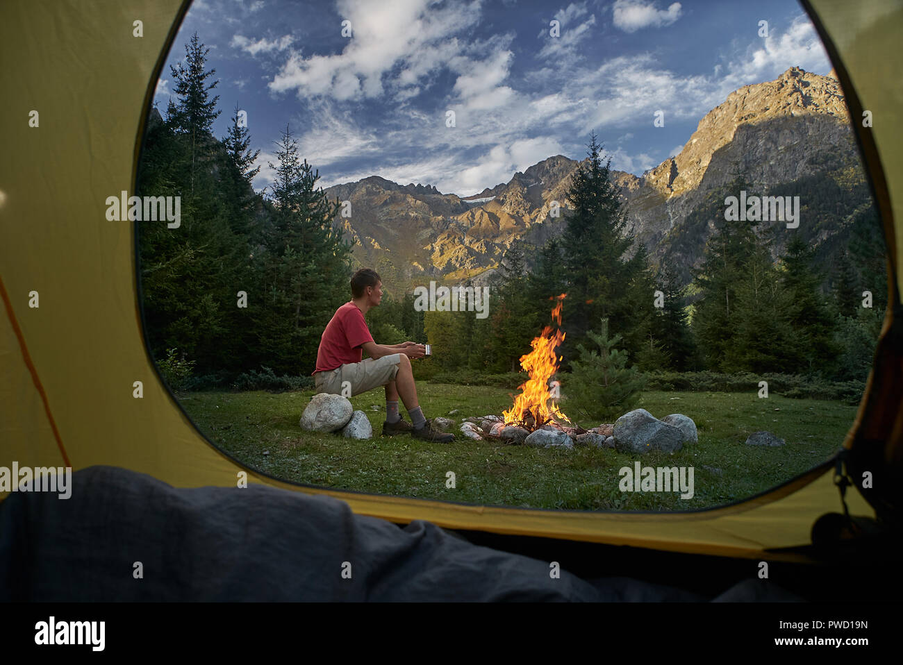 Vista dall'interno di turista tenda al tramonto. Da solo turista, l'uomo escursionista seduti al falò, tenendo in mano un bicchiere e guardando le montagne Foto Stock