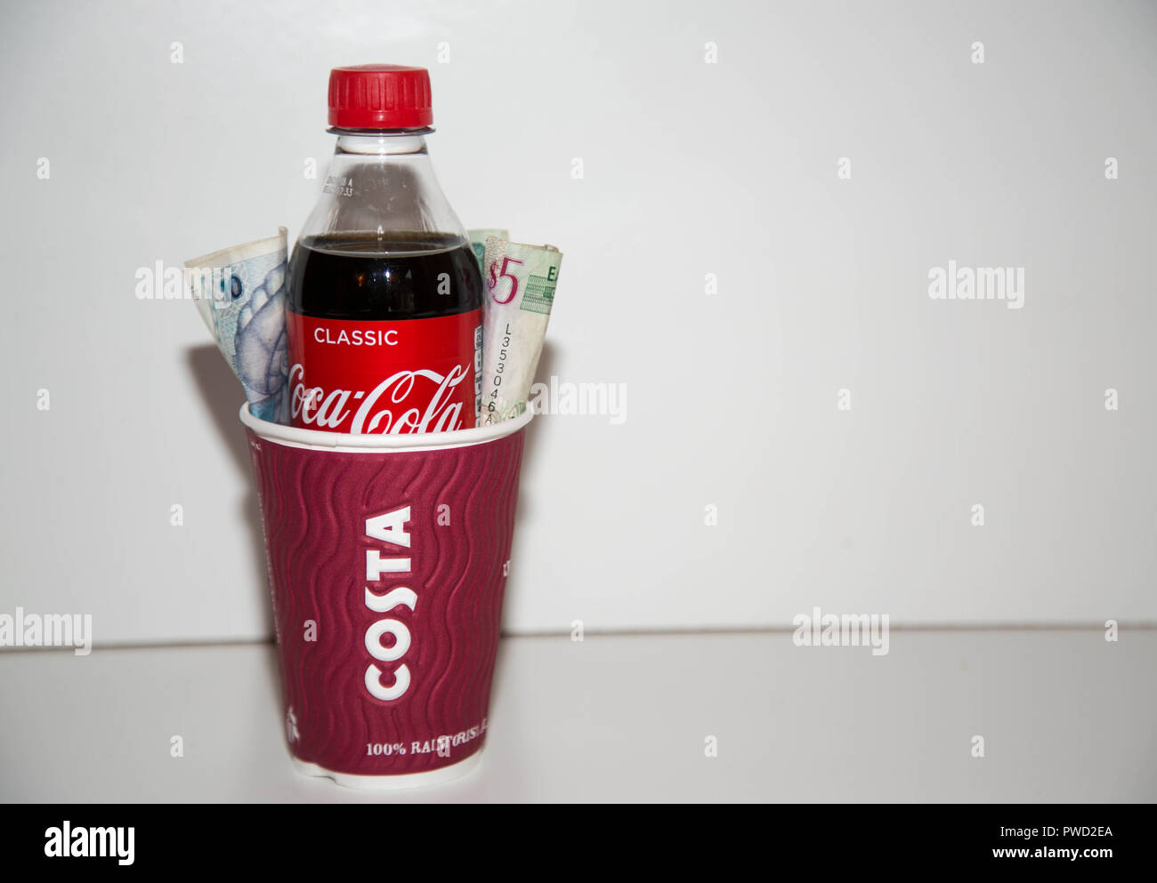 Costa Coffee Cup con coca cola bottiglia all'interno con contanti ogni lato su sfondo bianco. Foto Stock