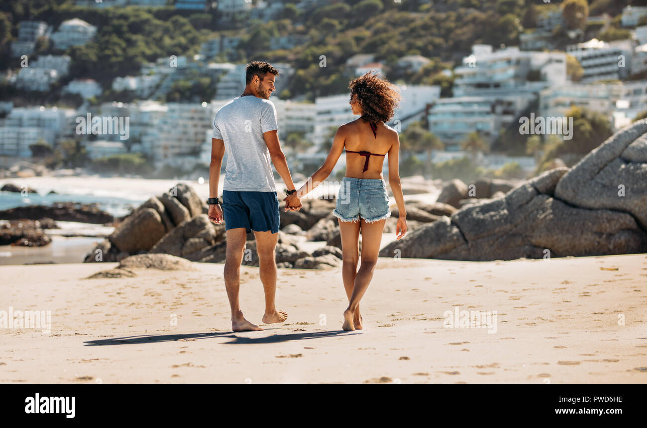 Turista giovane in vacanza a piedi sulla spiaggia guardando ogni altro. Vista posteriore di un paio di camminare su una spiaggia rocciosa e mano nella mano in una giornata di sole. Foto Stock