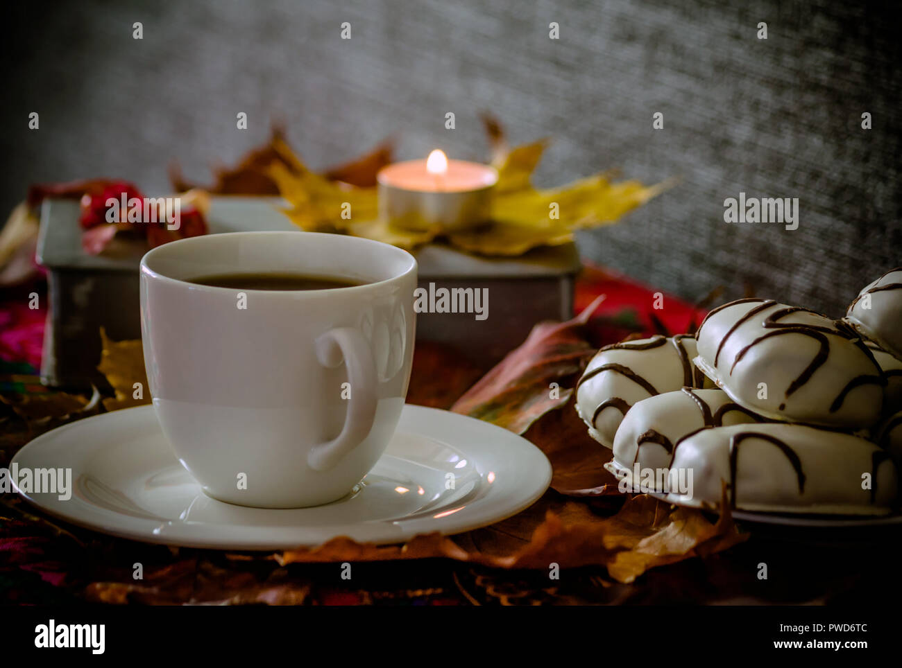 Tazza bianca con piattino con tè o caffè è sul tavolo, foglie di acero, accanto ad esso è una piastra con i cookie di bianco con strisce di cioccolato, un libro Foto Stock