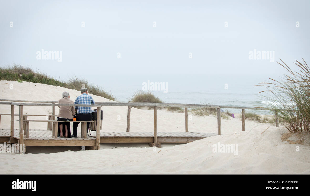 Costa Nova, Portogallo - Agosto 2018: Cute vecchia coppia seduta nel banco di legno guardando il mare in Costa Nova Beach. Foto Stock