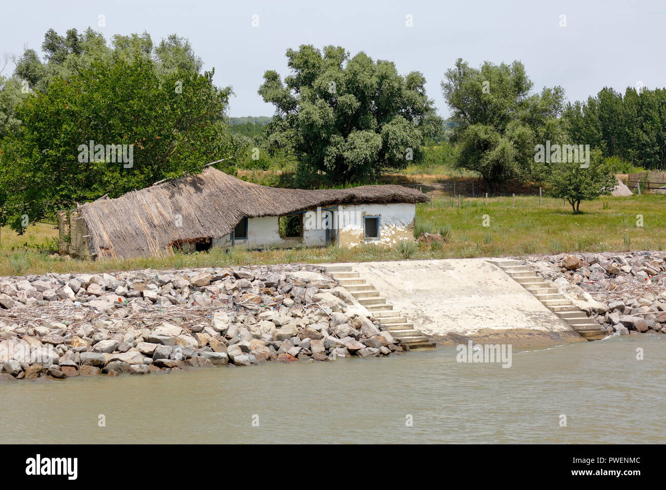 Cottage vicino Vulturu al ramo di Sulina del Danubio, imbarcadero, dalle rive del Danubio, waterside promenade, paesaggio fluviale, gruppo di alberi, Danubio dyke da massi, la protezione dalle inondazioni, palude, terreni agricoli, Romania, Tulcea contea, Dobrudja, il Delta del Danubio, Riserva della Biosfera del Delta del Danubio, sul delta del fiume, estuario del fiume Danubio in bocca al Mar Nero, Sito Patrimonio Mondiale dell'UNESCO, monumento naturale Foto Stock