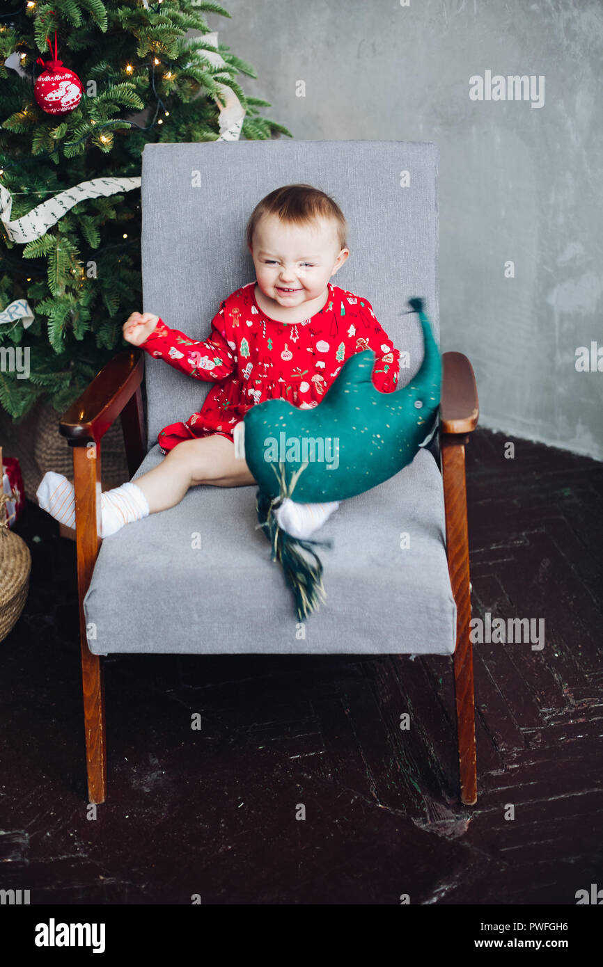 Adorable kid in abito rosso con stampa di natale seduto in poltrona. Foto Stock