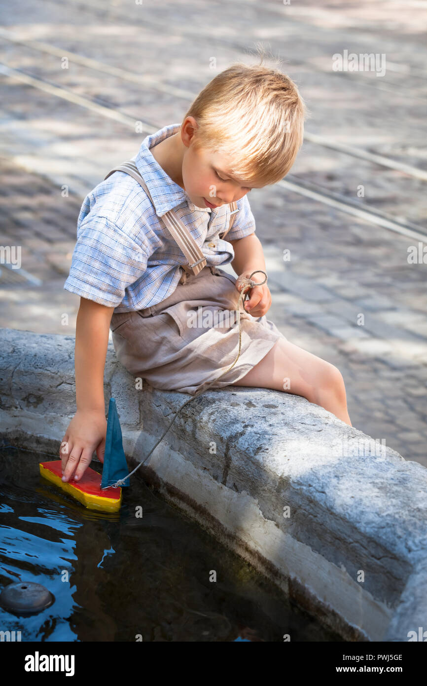 Scena nostalgico: cute little boy seduta sul bordo di piccole sassoso bacino idrico e giocare con la sua colorata giocattolo di legno barca Foto Stock