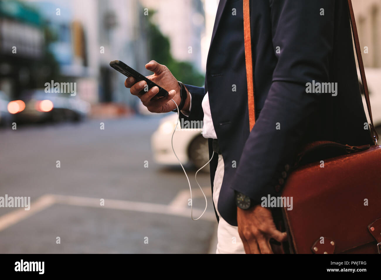Ritagliato shot di imprenditore passeggiate all'aperto utilizzando il telefono cellulare. Uomo in tuta con borsetta a camminare su una strada di città con il cellulare in mano. Foto Stock