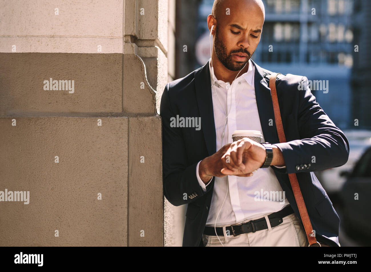 Un uomo nero serio e bello in abbigliamento formale che controlla il tempo mentre si appoggia a un muro lungo una strada della città. uomo d'affari che aspetta fuori dalla strada. Foto Stock