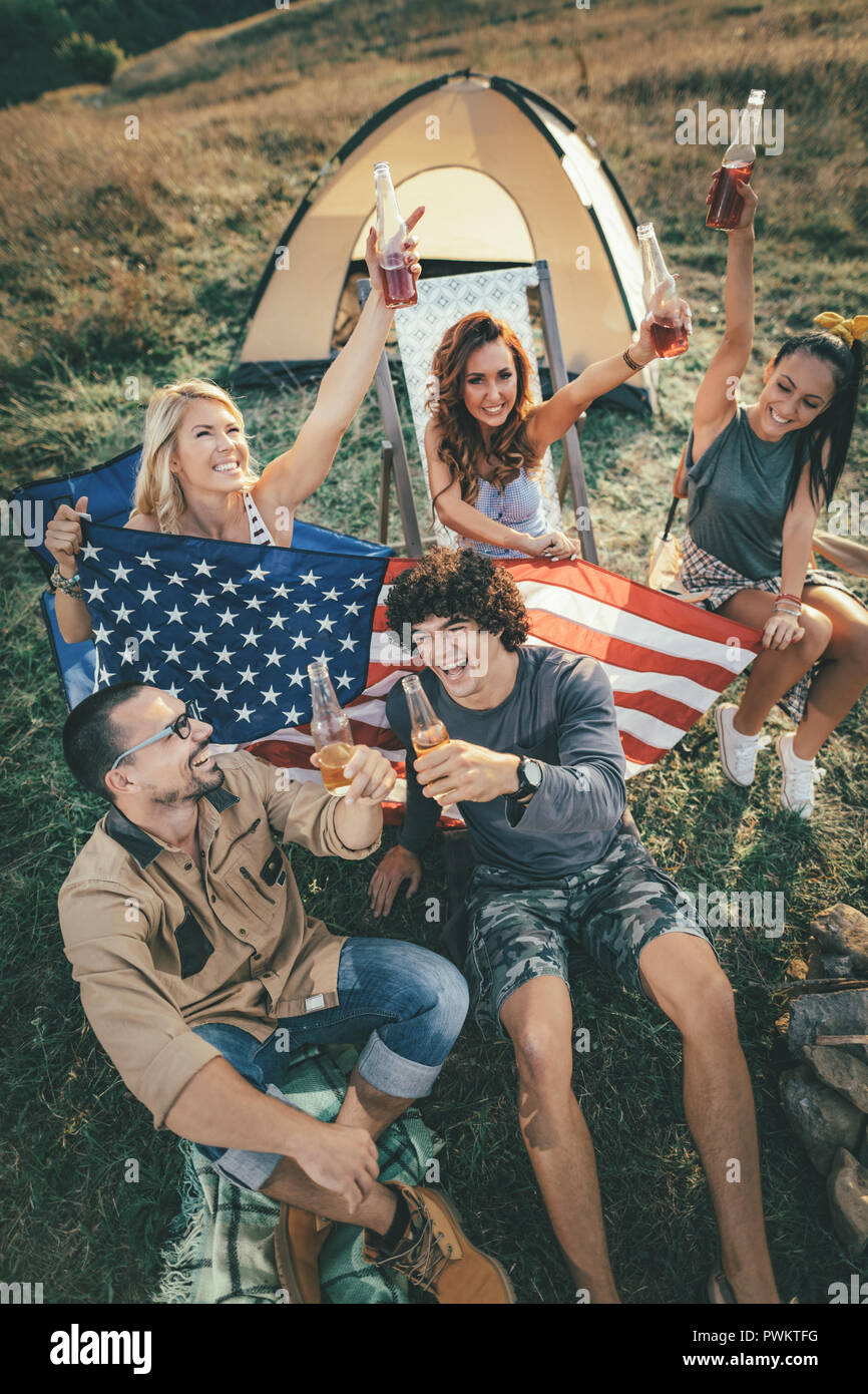 Felice giovani amici quarta di luglio di celebrazione e di godere di una giornata di sole in montagna. Si stanno tenendo bandiera americana, ridendo e tostatura con birra b Foto Stock