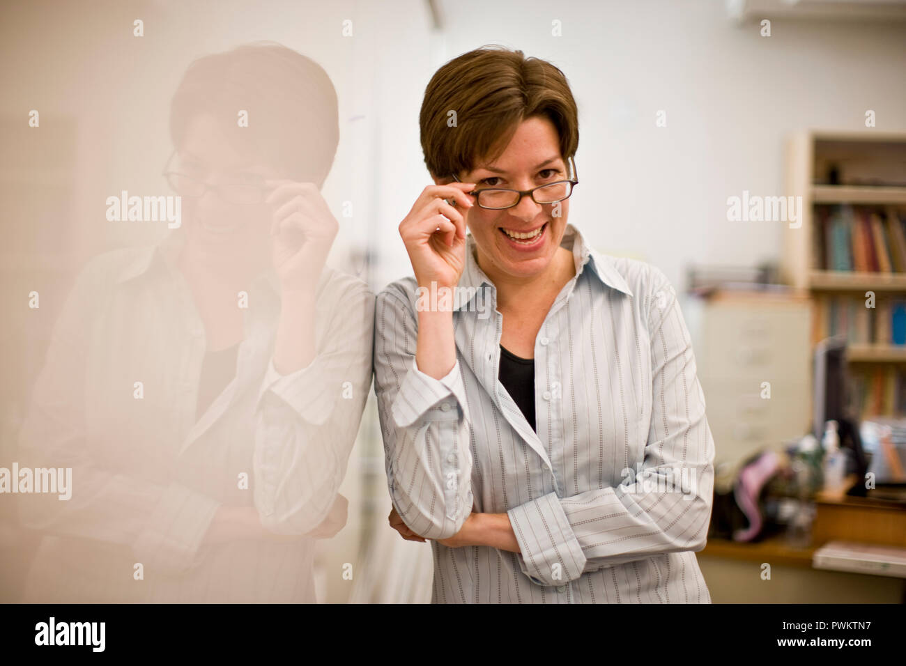 Ritratto di una donna sorridente insegnante adegua i suoi occhiali accanto a una lavagna in un'aula. Foto Stock