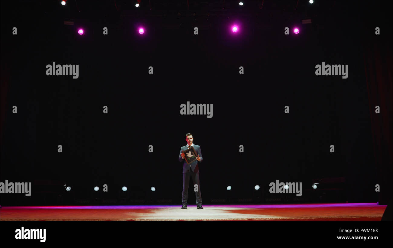 Showman. Giovane maschio intrattenitore, presenter o attore sulla scena. Schiena, braccia ai lati, fumo sullo sfondo di Spotlight. Vista posteriore di un maschio di oratore pubblico che parla al microfono, puntando in la. Foto Stock