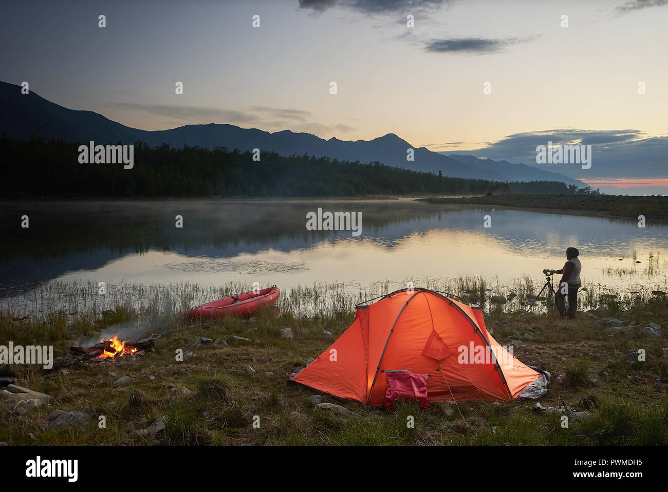 Fotografo è scattare una foto del bellissimo lago e montagna magica con la luce del tramonto. Camping con tenda arancione, canoa e falò. Foto Stock