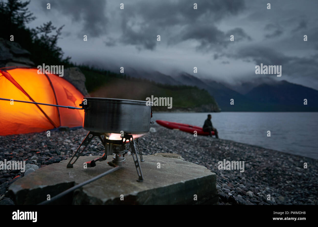 La preparazione del caffè e del tè con un fornello a gas presso il campeggio con rosso illuminato tendono. Vista del lago a nuvoloso notte. Sfocato silhouette di un uomo sullo sfondo. Foto Stock