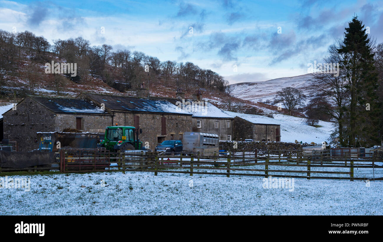 Il trattore e i rimorchi da edifici agricoli e la vecchia casa colonica su terreni innevati giornata invernale in scenic, Yorkshire Dales campagna valley - Hubberholme, Inghilterra, Regno Unito. Foto Stock