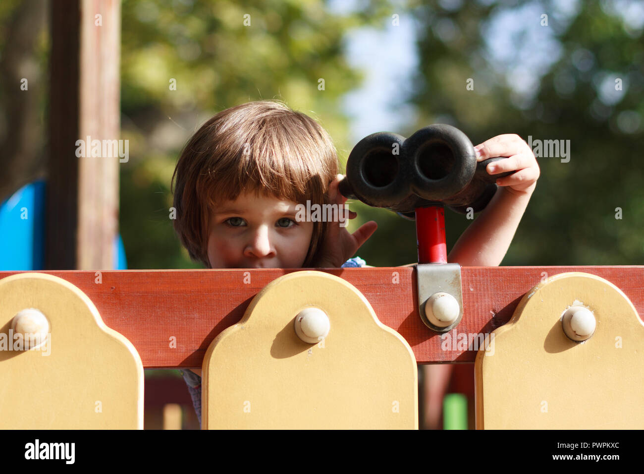 Bambino curioso che gioca con un telescopio giocattolo in un parco giochi che sbircia su attrezzature colorate Foto Stock