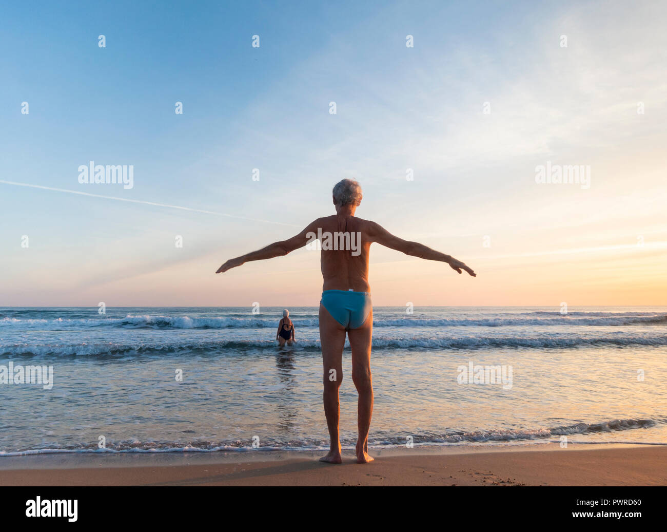Uomo anziano stretching prima di nuotare sulla spiaggia a sunrise. Foto Stock