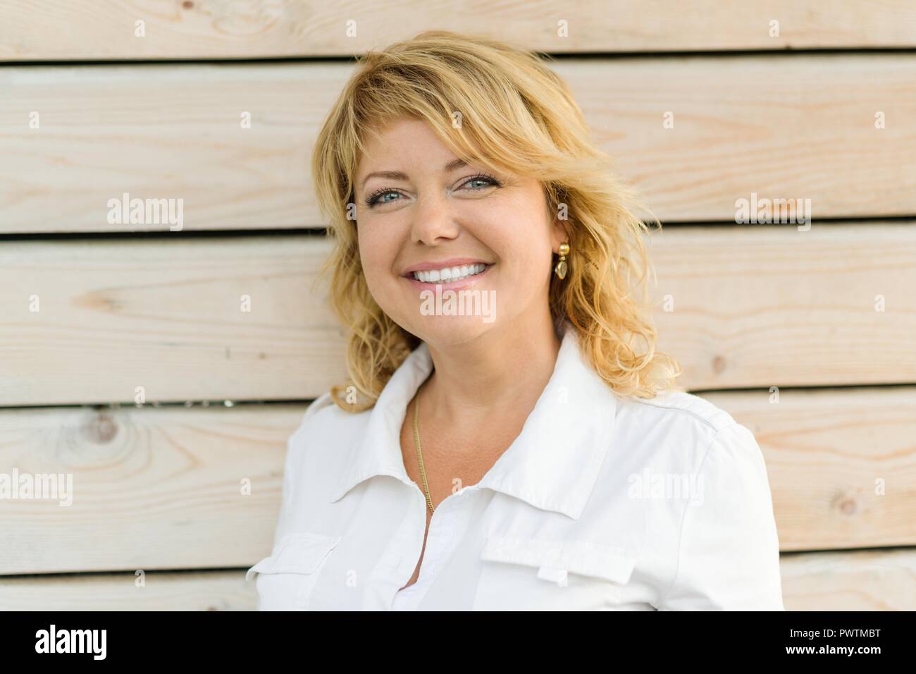 Ritratto maturo positiva di mezza età donna sorridente close-up. Donna bionda, all'aperto sullo sfondo di legno. Foto Stock