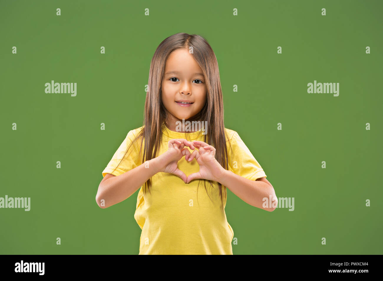 Bella sorridente ragazza teen rende la forma di un cuore con le mani sullo sfondo verde. Gesto di amore da piuttosto giovane bambino. Foto Stock