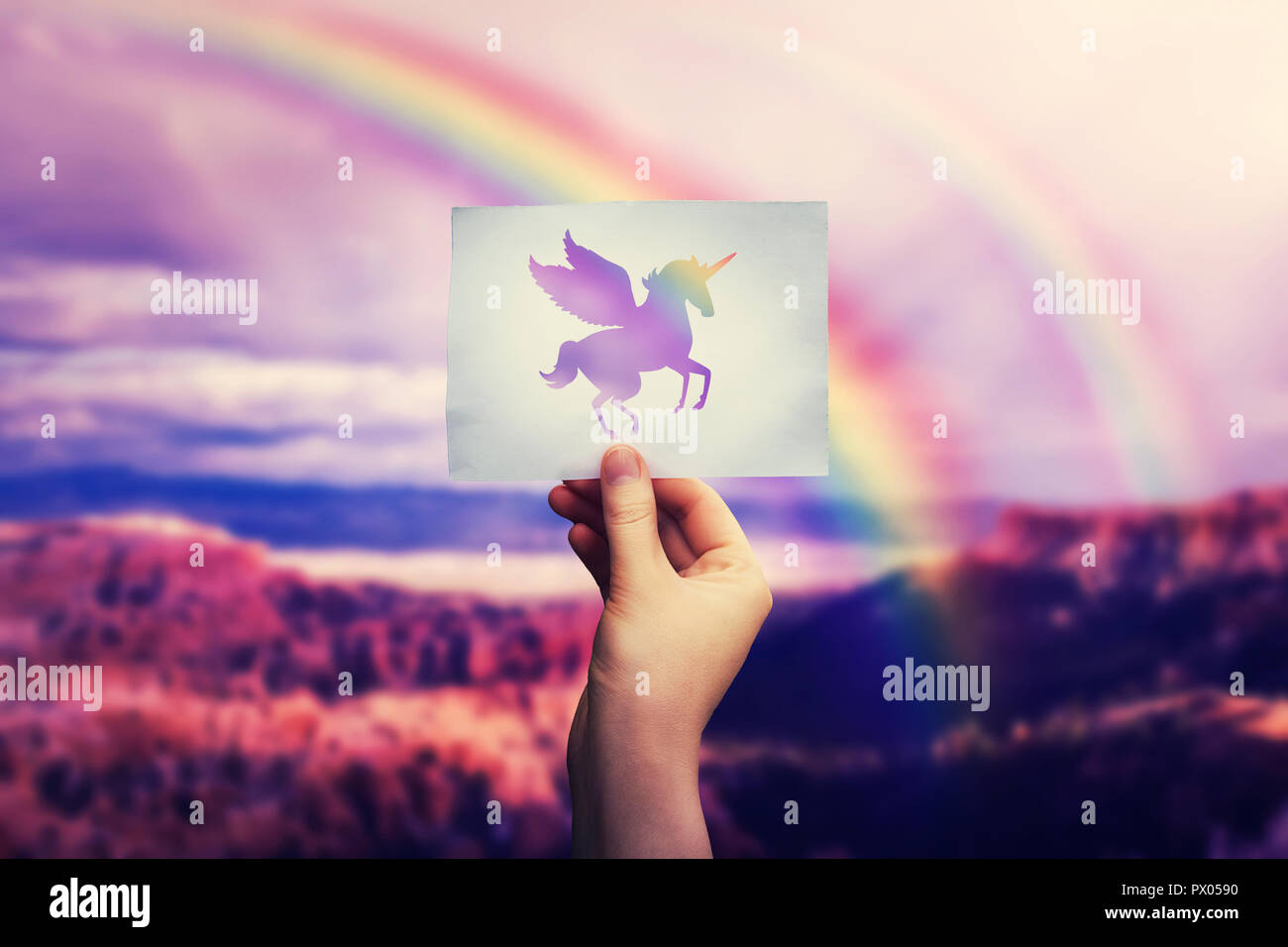 Donna mano tenendo un foglio di carta con pegasus icona Unicorn sulla natura selvaggia valle sfondo su l'arcobaleno. Viaggio di amore e di libertà concetto, cont Foto Stock
