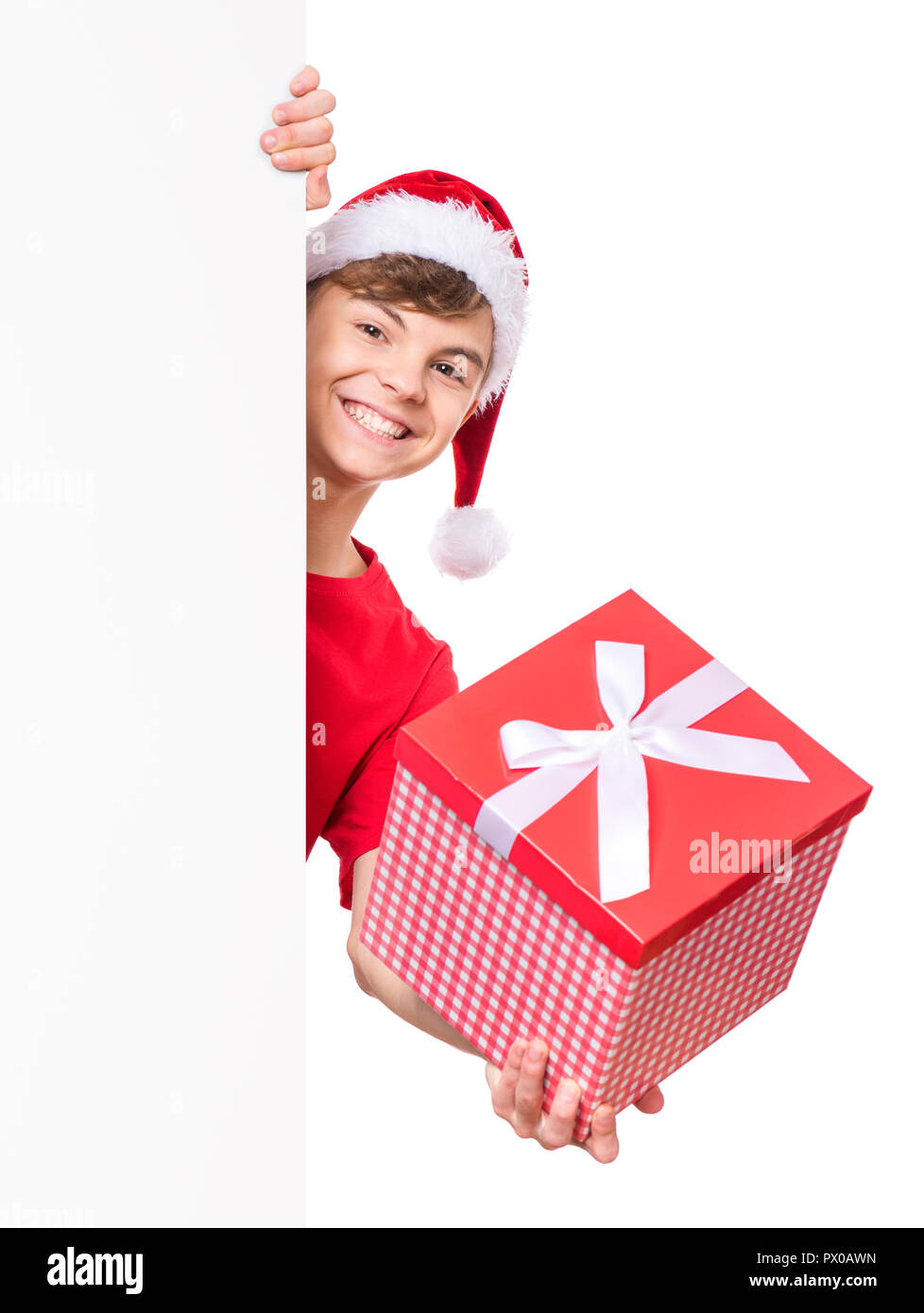 Ritratto di sorridere teen ragazzo con cappello di Natale. Adolescente azienda confezione regalo e sbirciare dietro vuoto vacanze a bordo. Felice carino bambino in posa dietro whi Foto Stock