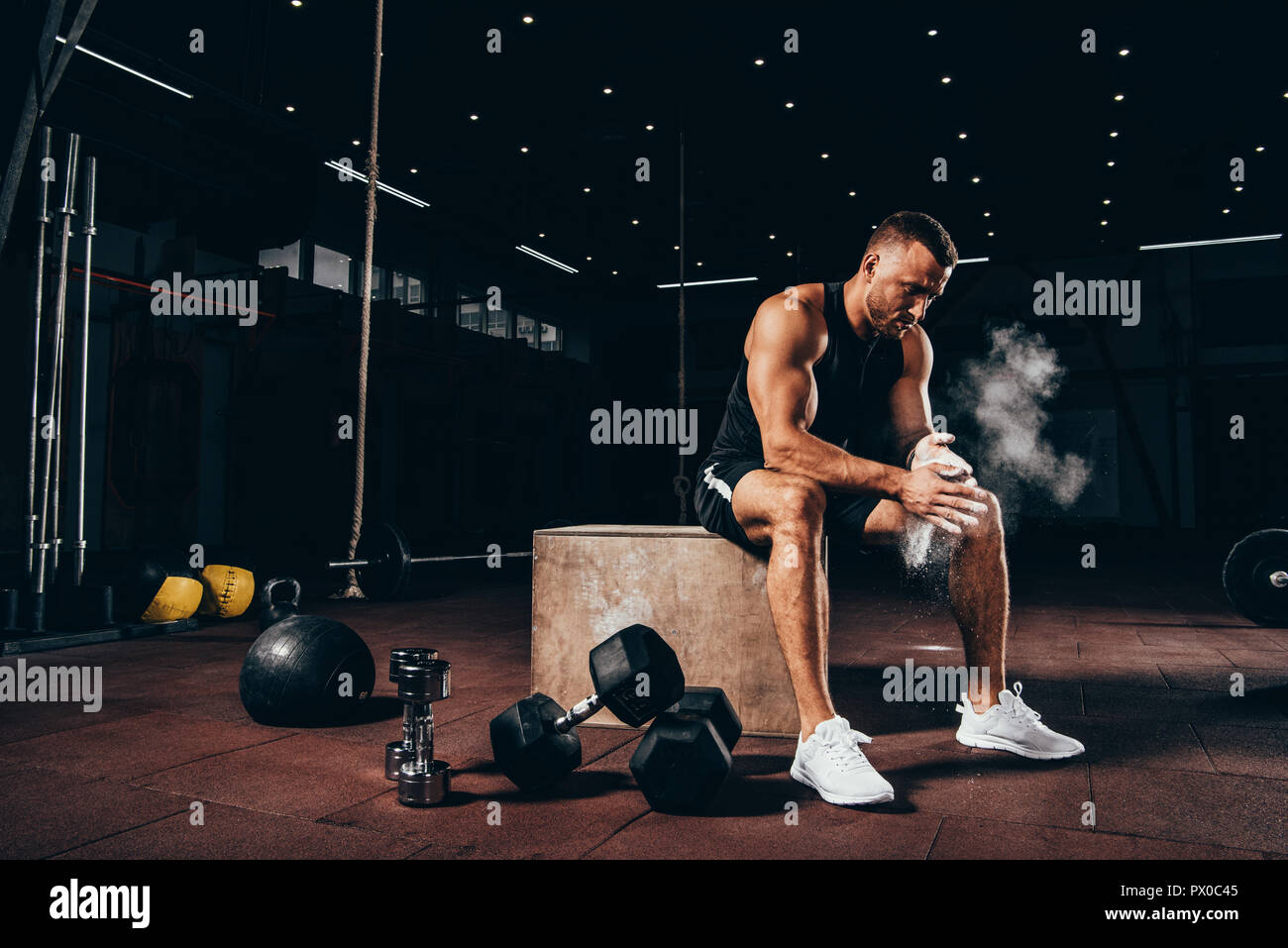 Bello athletic uomo seduto sul cubo con attrezzature da palestra intorno e battendo le mani con talco prima di allenamento Foto Stock