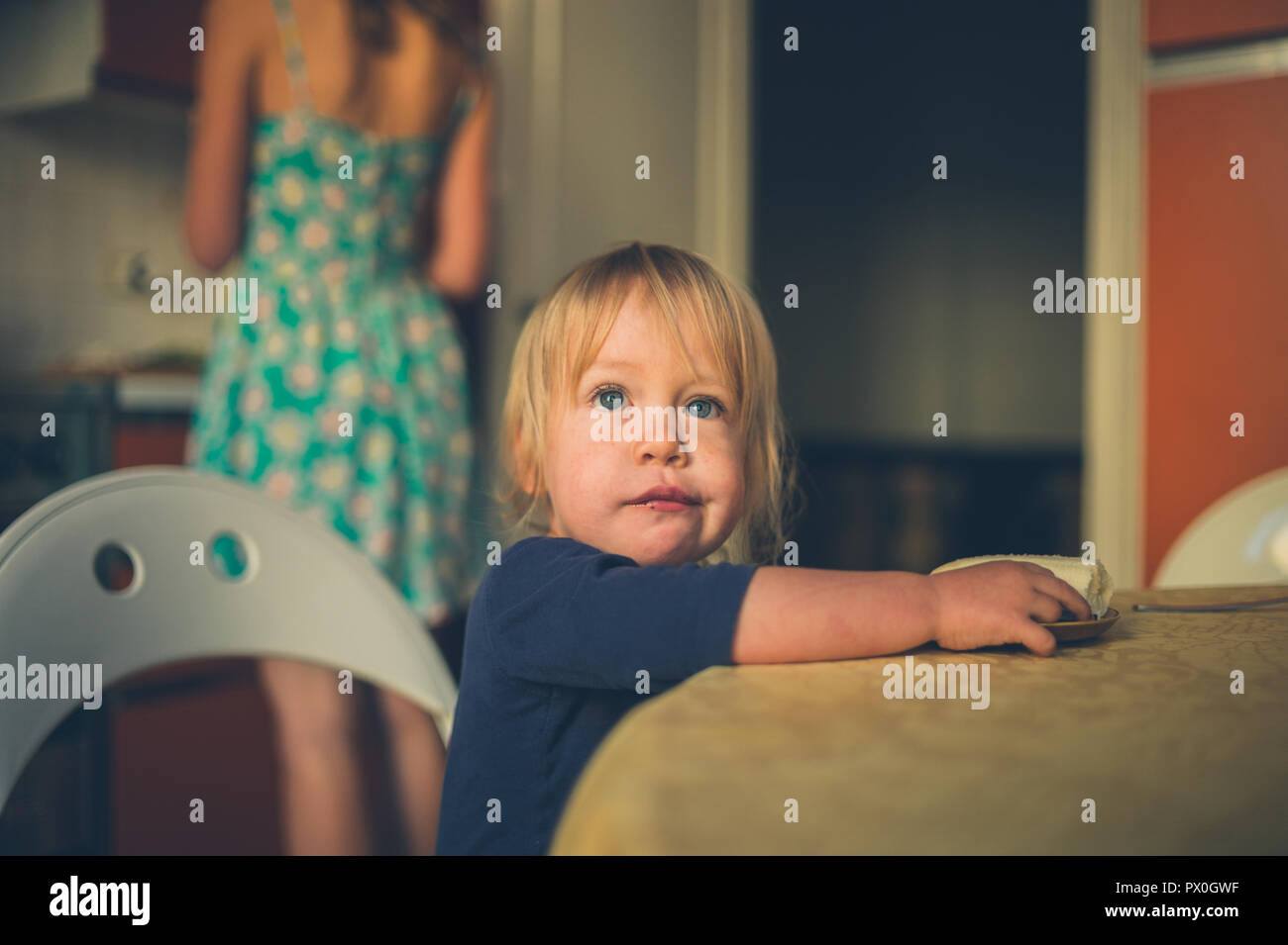 Un piccolo bambino è seduta in cucina con sua madre la cottura in background Foto Stock