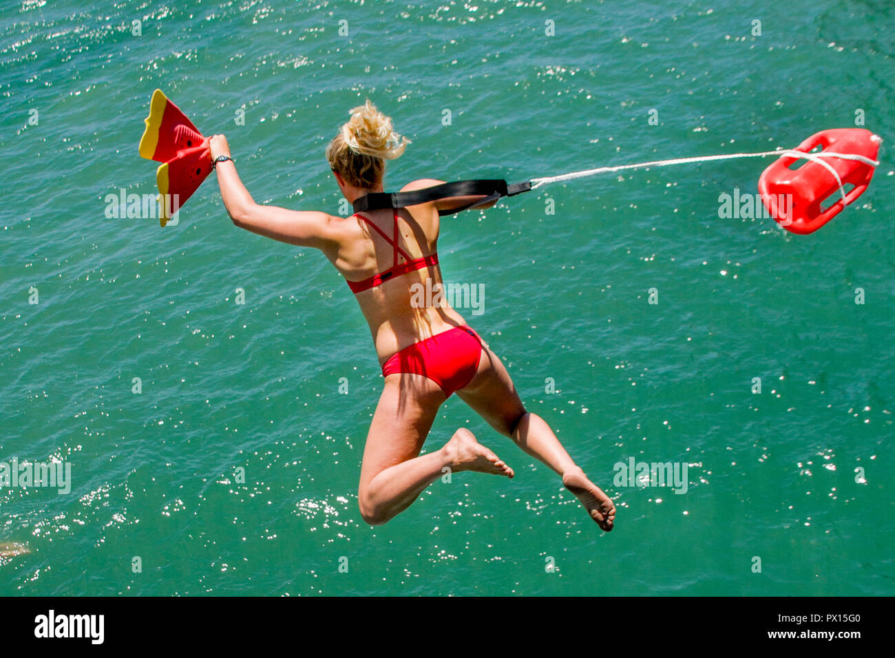 Portante galleggiante e pinne da nuoto, una giovane donna adulta bagnino di salvataggio in un bikini Rosso si prepara a saltare da un molo di Huntington Beach, CA, per il salvataggio di una vittima di annegamento. Foto Stock