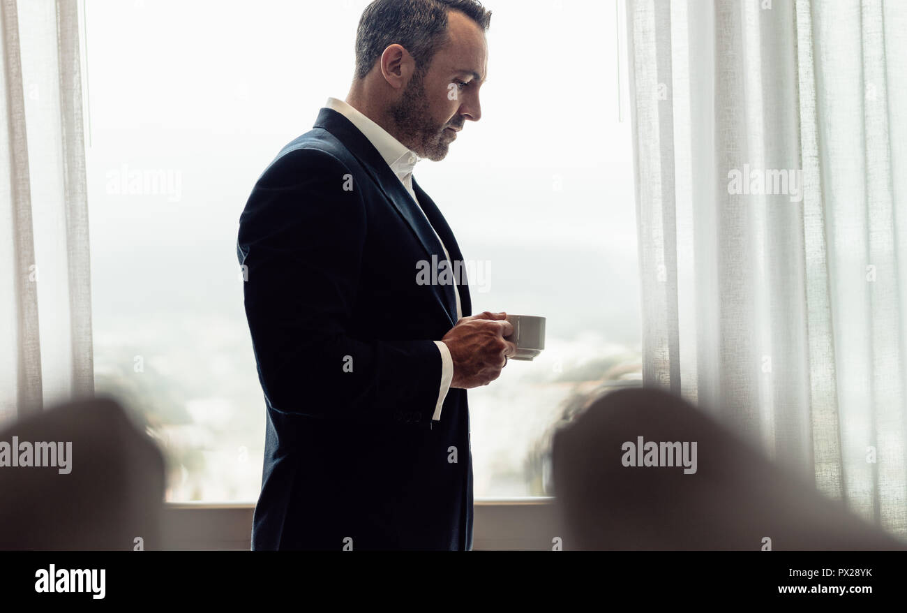 Imprenditore in piedi in camera in hotel con una tazza di caffè. Uomo maturo in tuta di bere il caffè in camera di albergo in mattina. Foto Stock