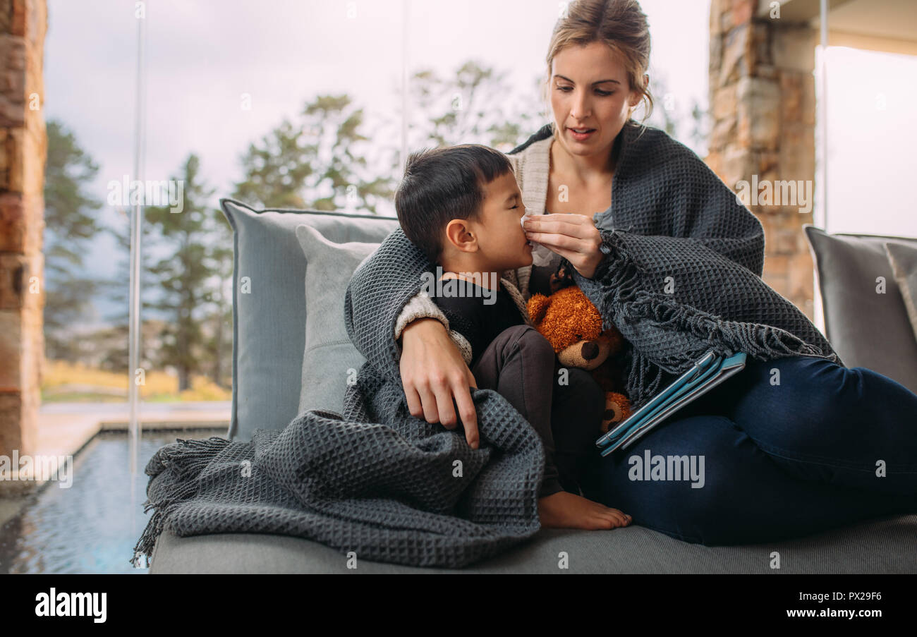 Madre tergi naso del suo piccolo figlio. Madre e figlio seduti sul divano avvolto in una coperta con una tavoletta digitale. La madre sta aiutando il suo figlio soffiare il naso Foto Stock