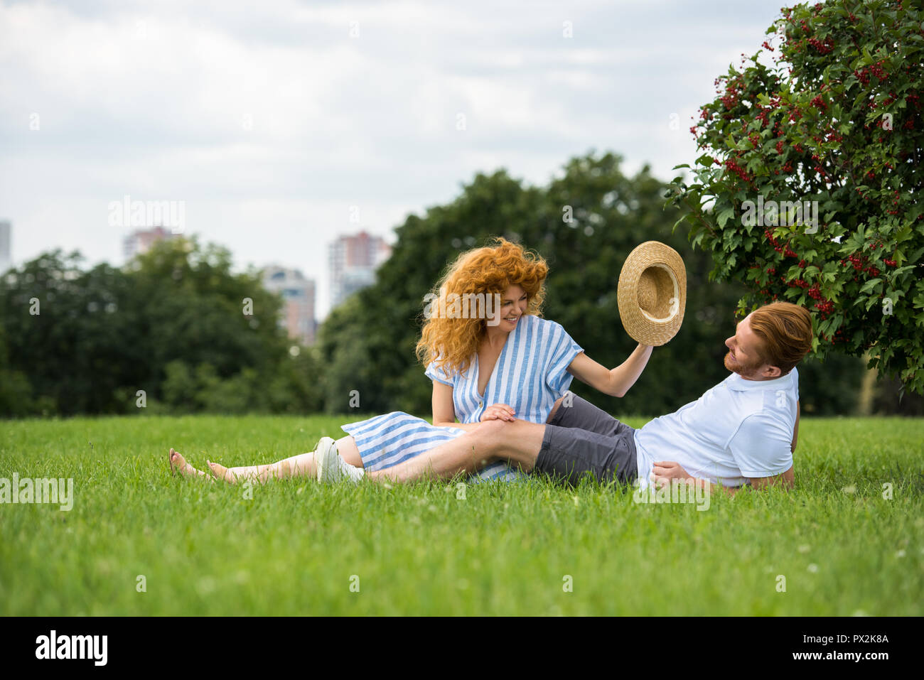 Sorridente redhead donna cercando di mettere il proprio cappello di paglia sulla testa di ragazzo di erba in posizione di parcheggio Foto Stock